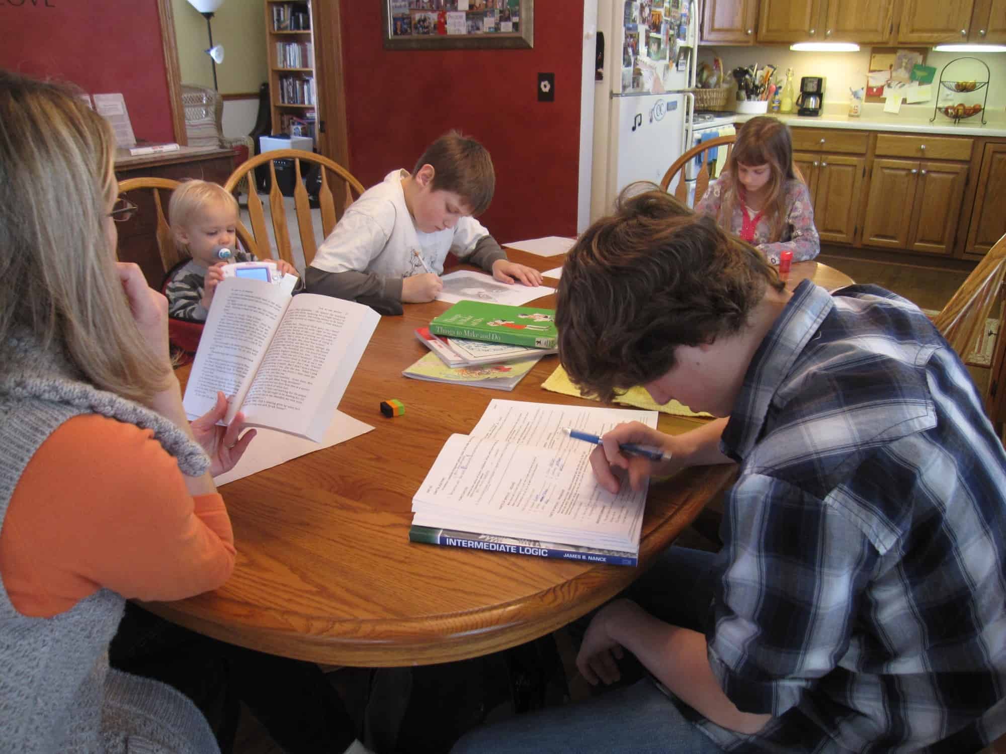 students sitting around the dining room table working on their classes. Mom is reading.