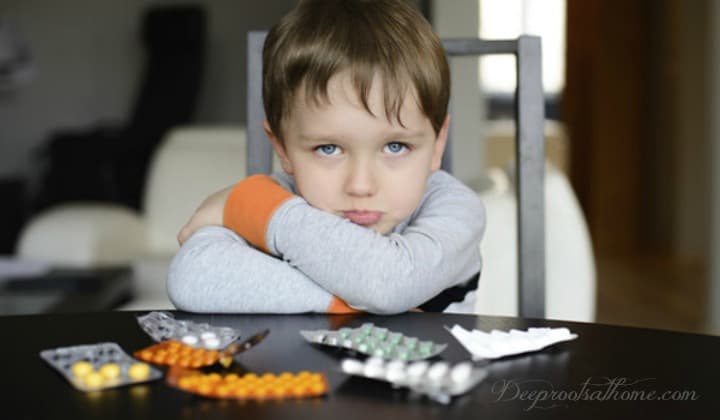 8M US Kids Taking Psychiatric Drugs: Parents Not Told The Deadly Documented Risks. A pouting boy at a table with colorful psychiatric drugs in their packages spread out before him.