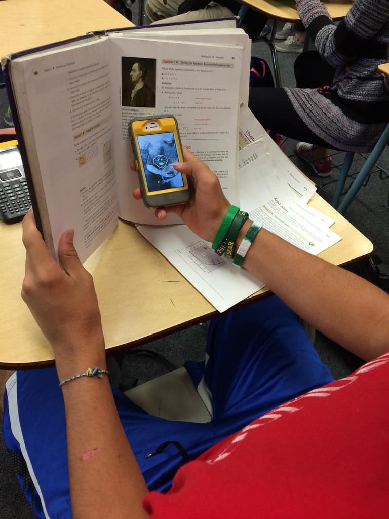 7 Truths Parents & Adults Don't Get About Today's High School. A student hiding his game on the iPhone behind his school book.